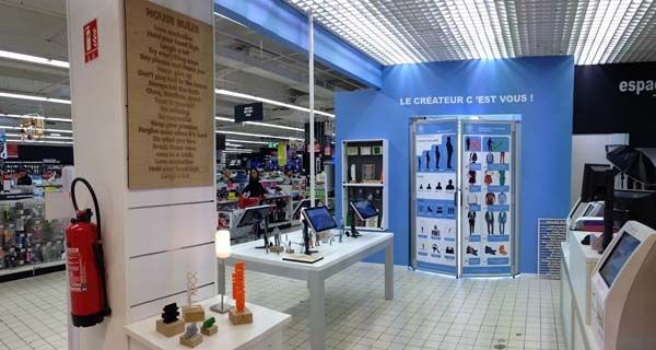 Reparto stampe 3D Auchan: stampanti 3D, estintore rosso, espositori con modelli. Innovazione e tecnologia.